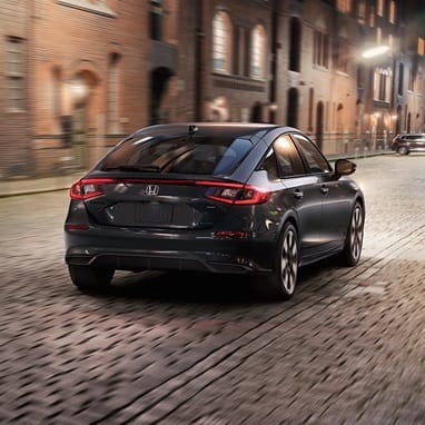 2027 Honda Civic Hatchback Sport Touring Hybrid in Meteorite Gray Metallic back driving on a cobblestone city street (Compact Hatchback).